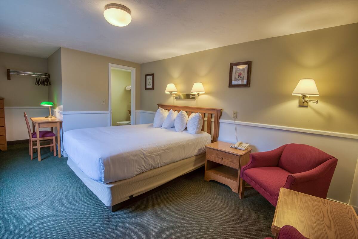 A hotel room with a bed, two bedside lamps, a red armchair, a desk with a lamp, and a chair.