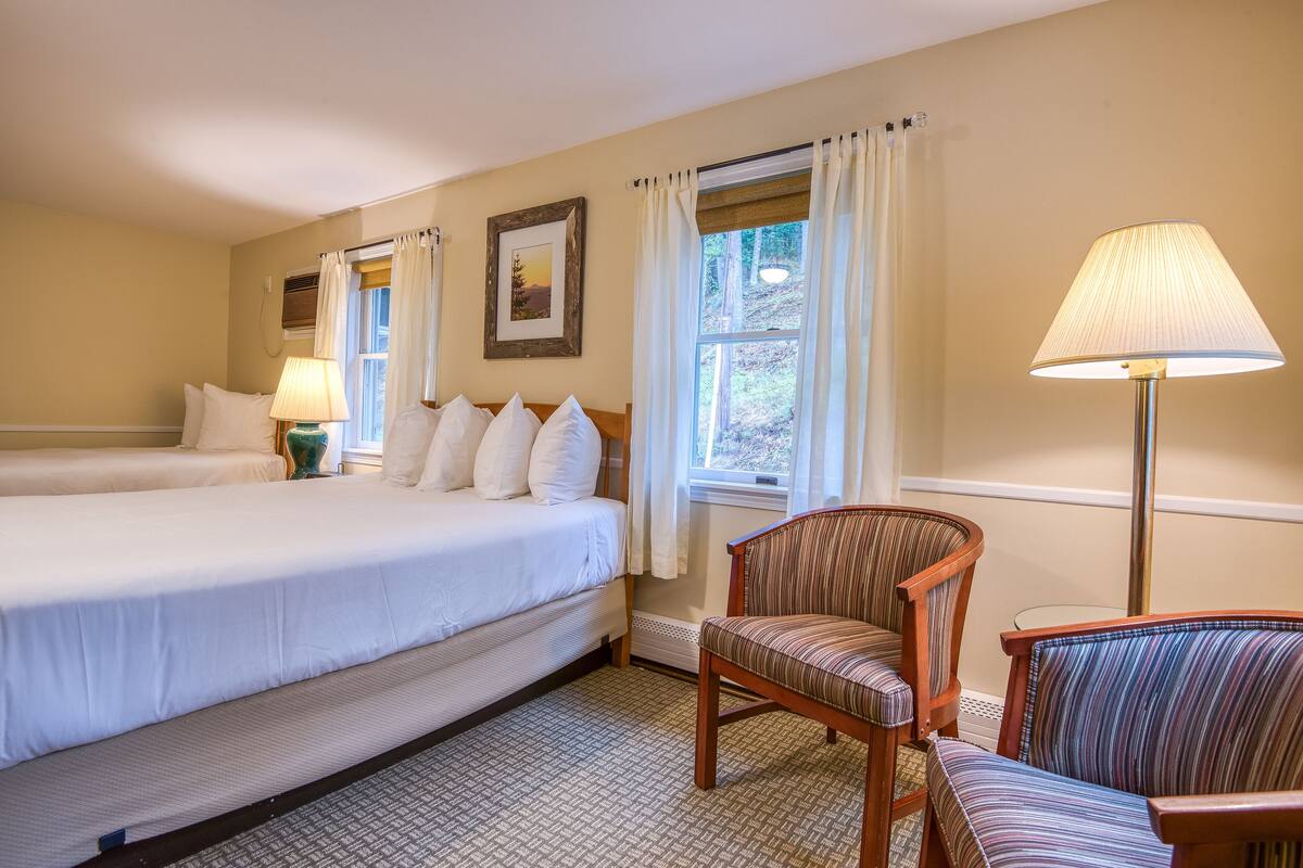 A hotel room with two beds, a chair, a lamp, and a window with curtains.