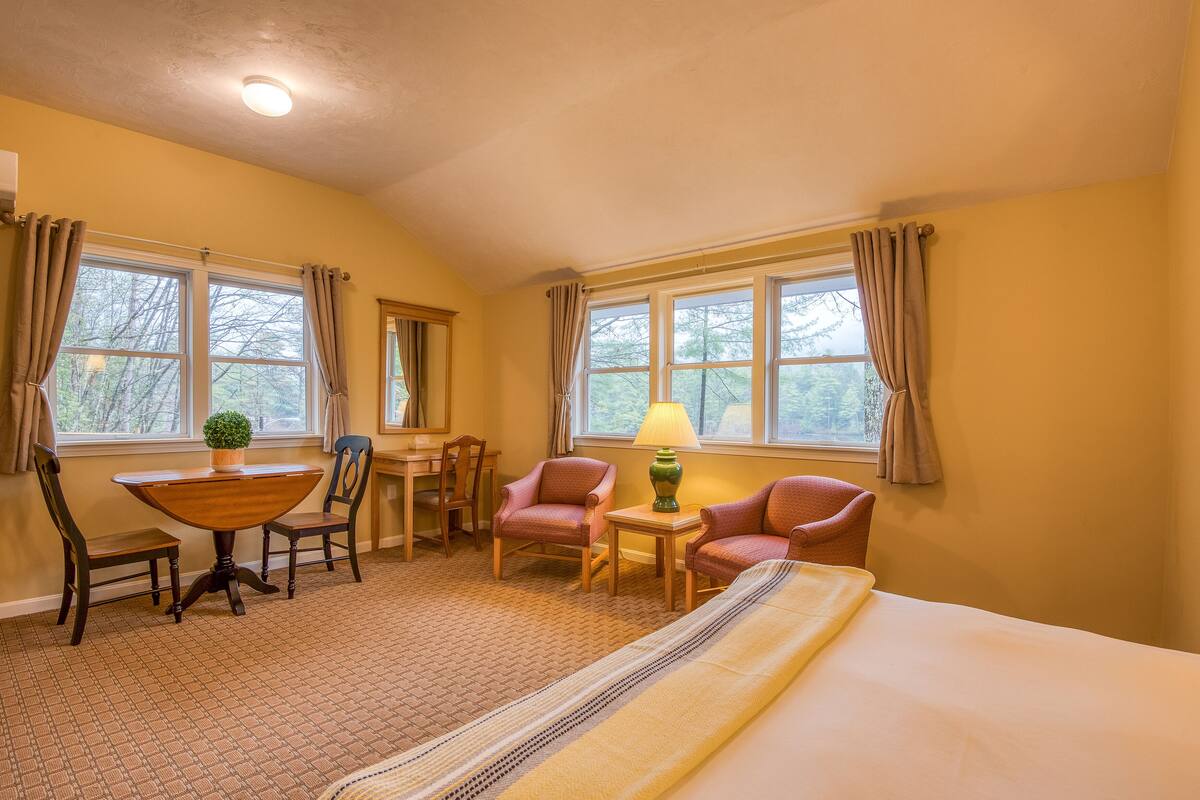 A hotel room with a bed, two chairs, a small table with a lamp, and two windows with curtains.