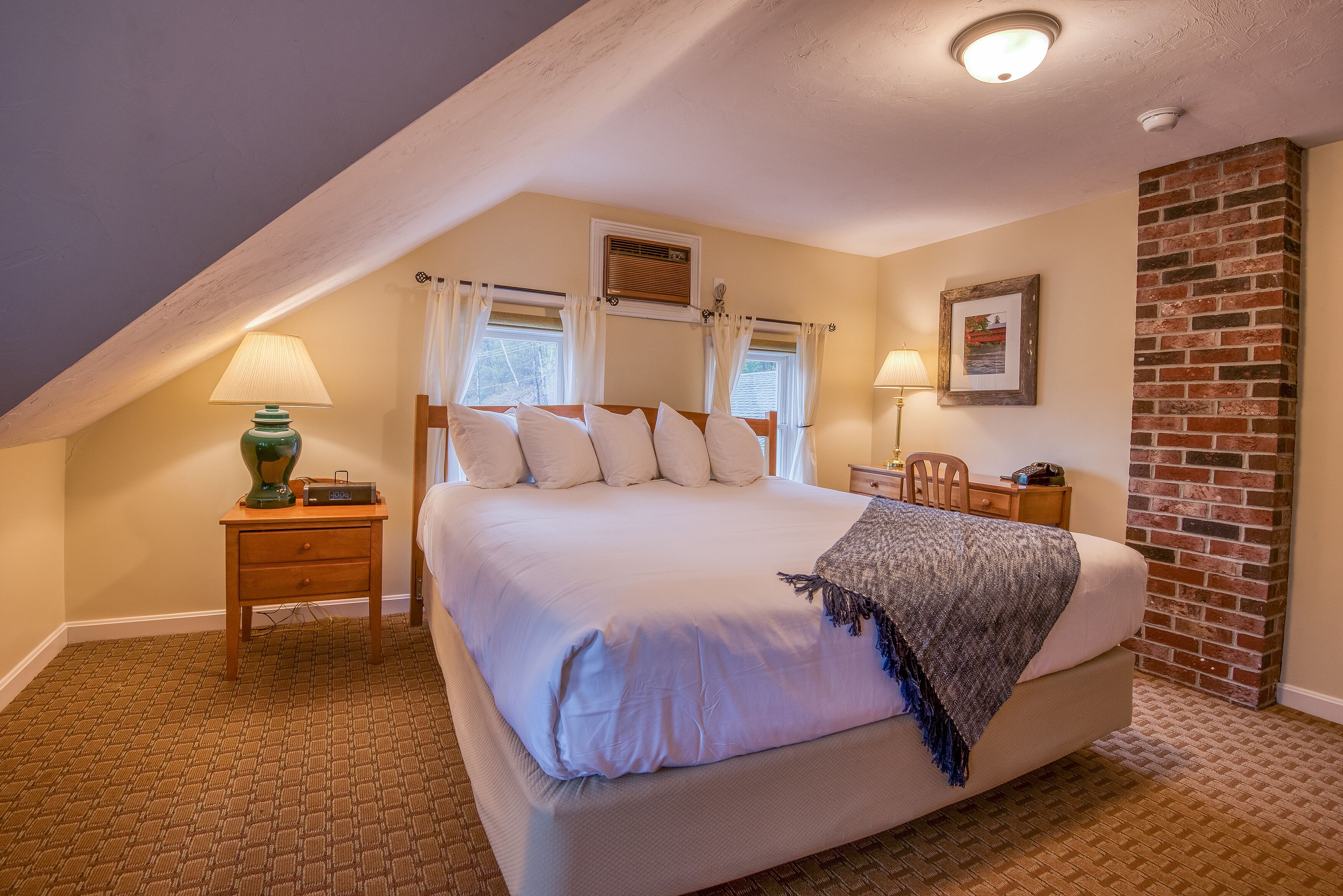 A bedroom with a large bed, two bedside tables, a lamp, a window with curtains, a picture frame, and a brick wall.