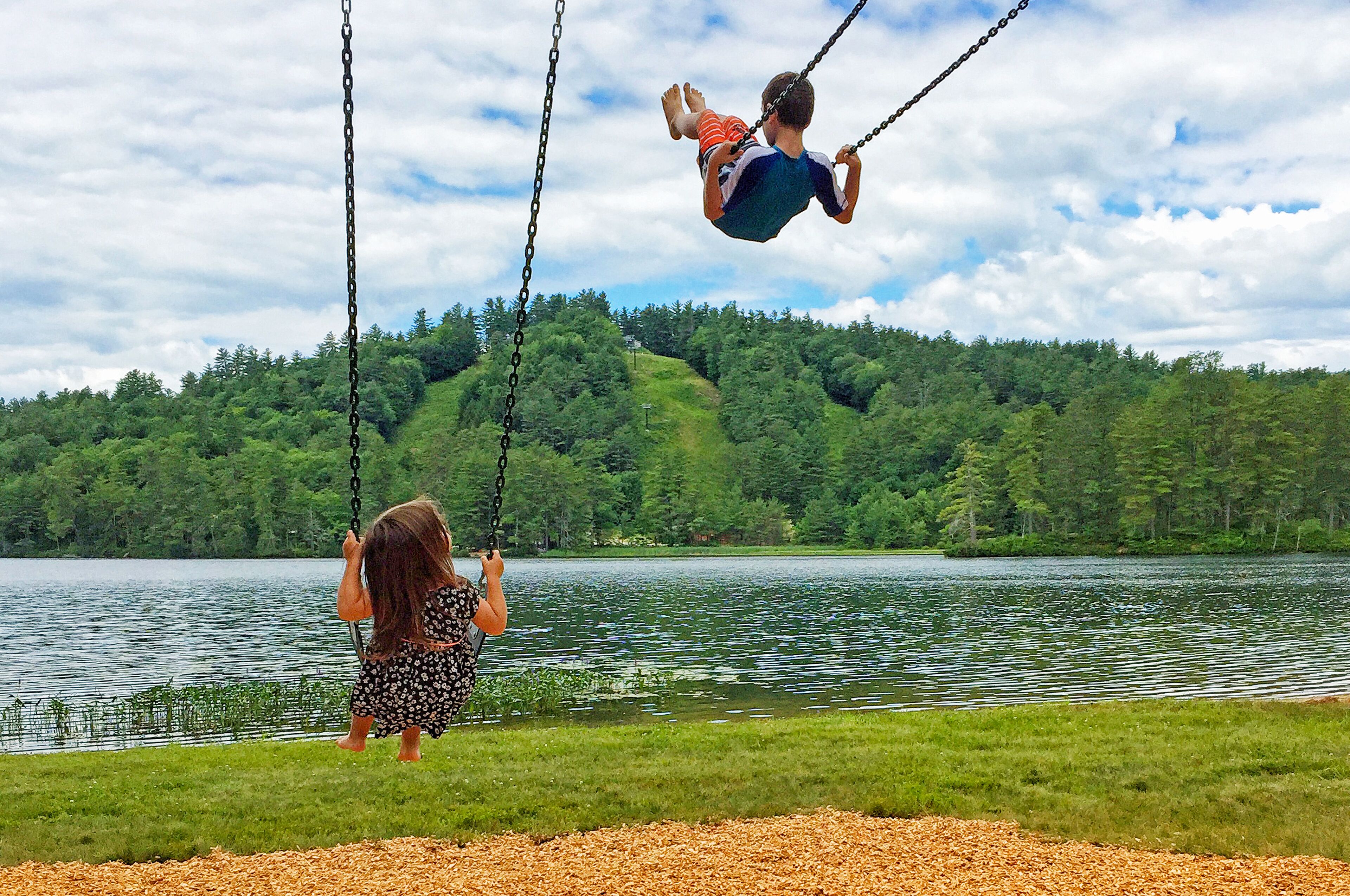 Two people on a swing over a lake with a forested hill in the background.
