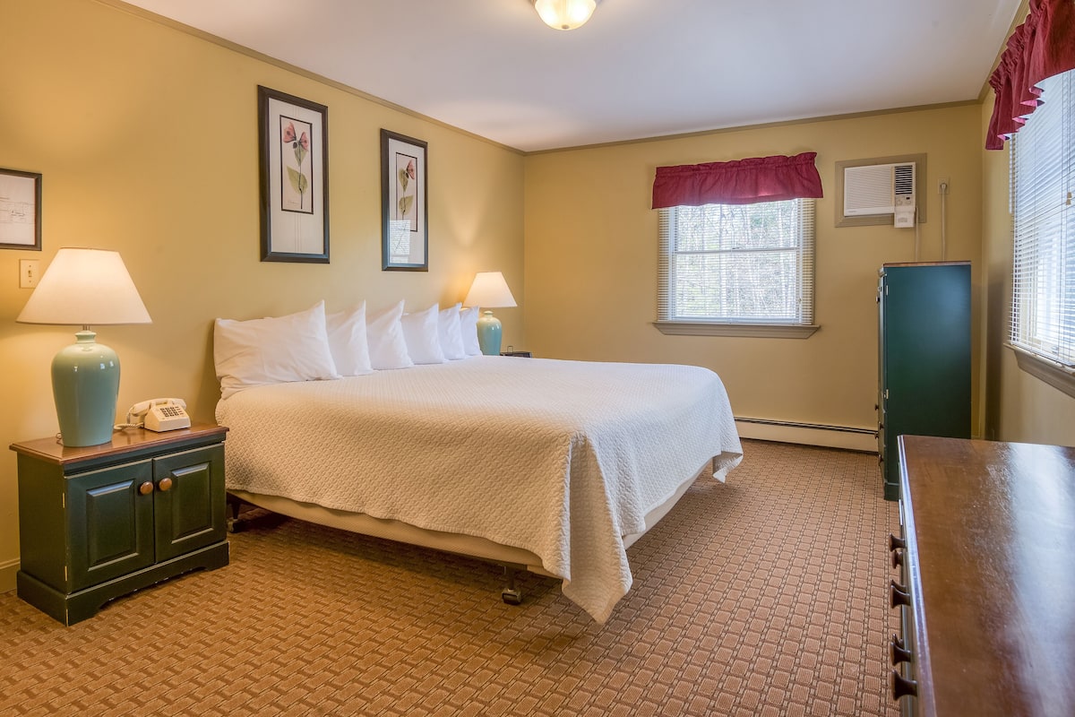 A neatly arranged bedroom with a bed, bedside lamps, a nightstand, a dresser, and a window with blinds.