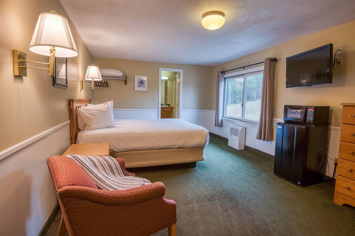 A hotel room with a bed, a chair, a desk, a TV, and a refrigerator.