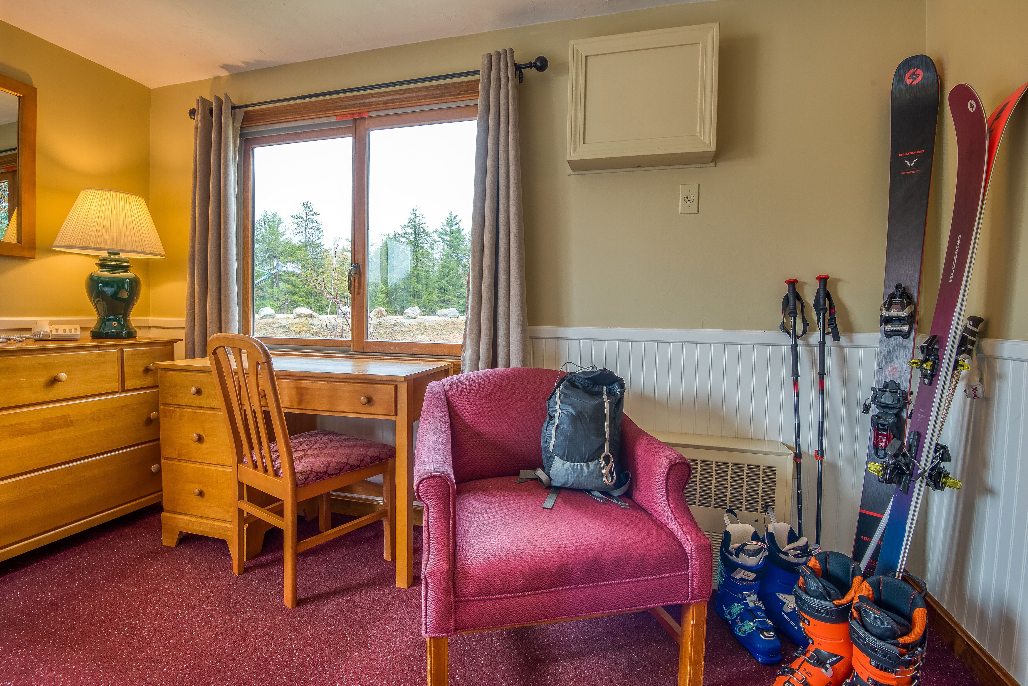 A room with a wooden desk, a chair, a lamp, a window with curtains, and skis leaning against the wall.