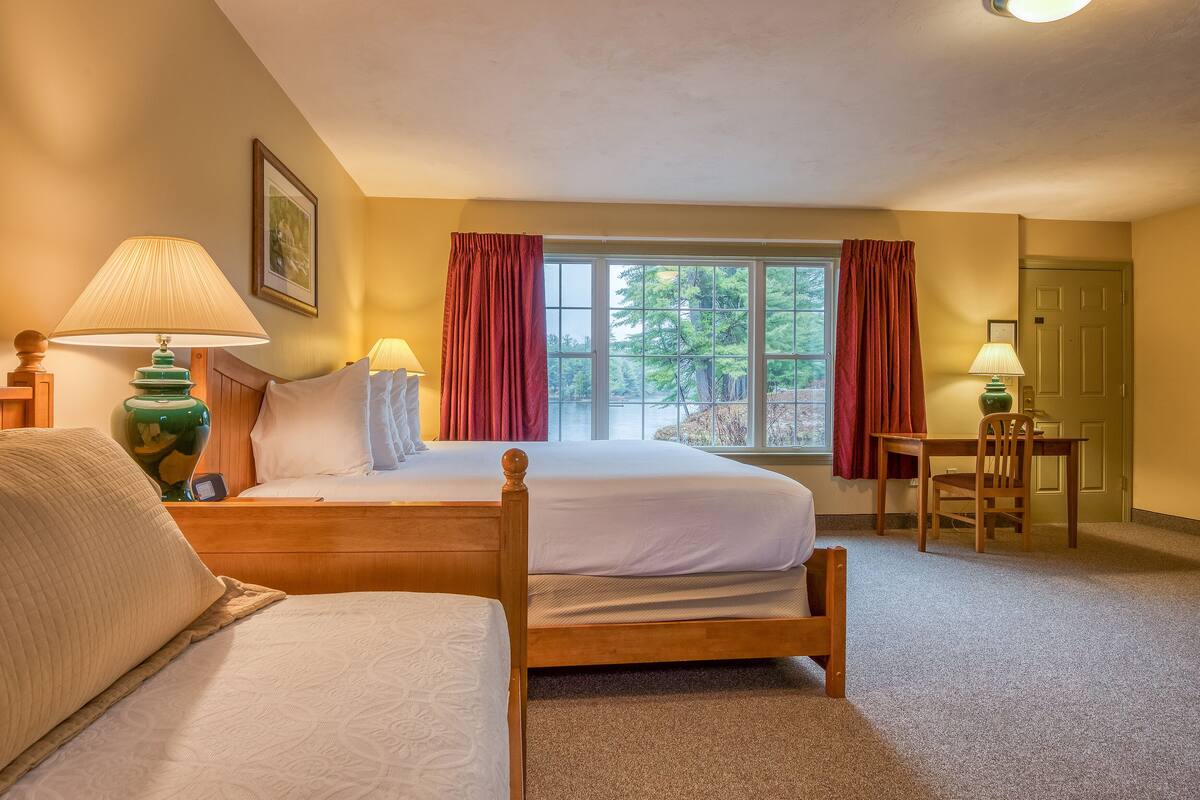 A hotel room with a large bed, two bedside tables with lamps, a window with curtains, and a door.