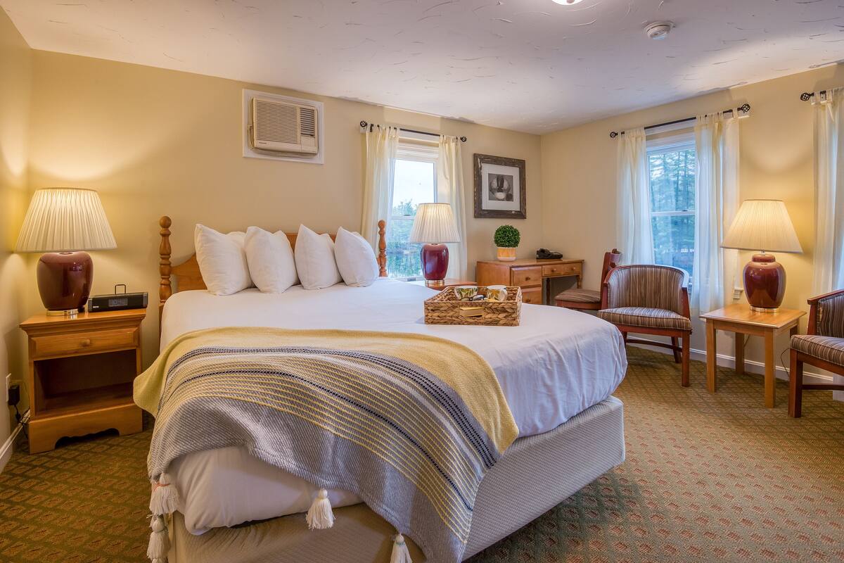 A neatly arranged bedroom with a large bed, bedside lamps, a small table with a lamp, a chair, and a window with curtains.