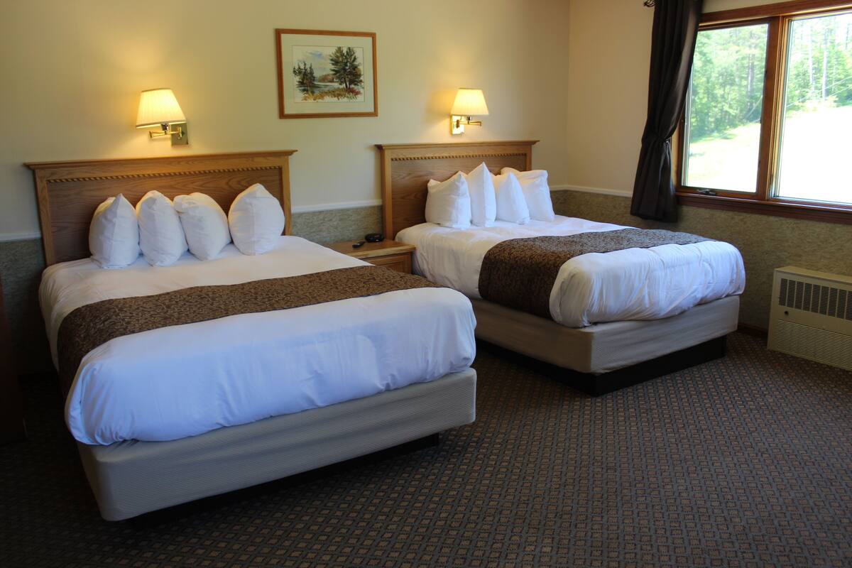 A hotel room with two beds, each with white linens and pillows, a headboard, bedside lamps, a framed picture on the wall, and a window with curtains.