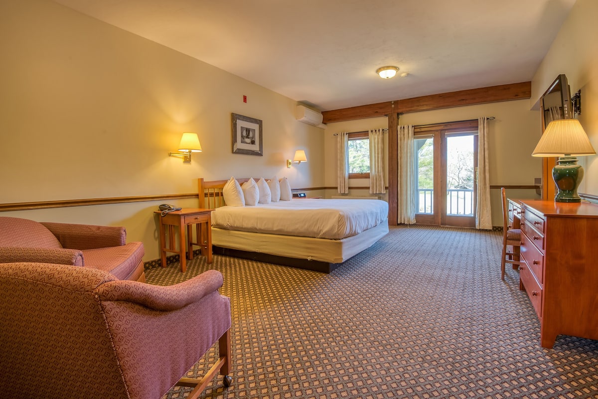 A hotel room with a large bed, two chairs, a desk, a dresser, and a window with curtains.