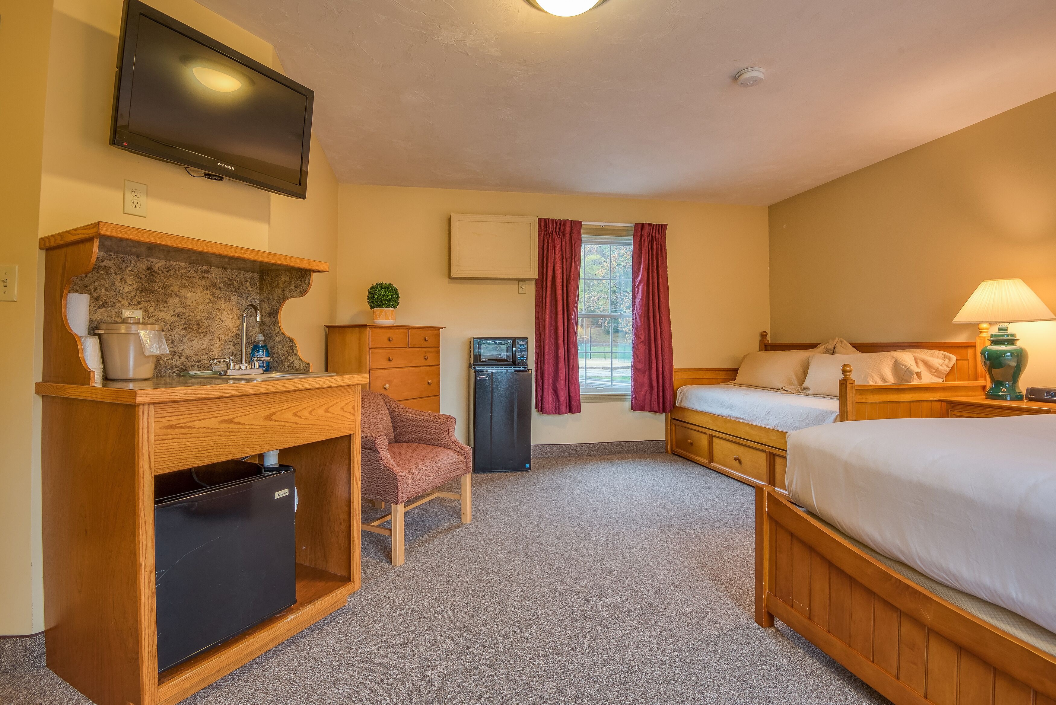 Quarto de hotel com escrivaninha de madeira, mini-frigorífico, televisão na parede, cama com roupa de cama branca e janela com cortinas.