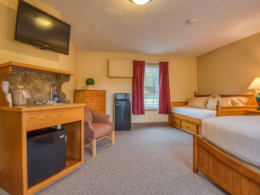 Habitación de hotel con escritorio de madera, refrigerador pequeño, televisor montado en la pared, cama con ropa blanca y ventana con cortinas.