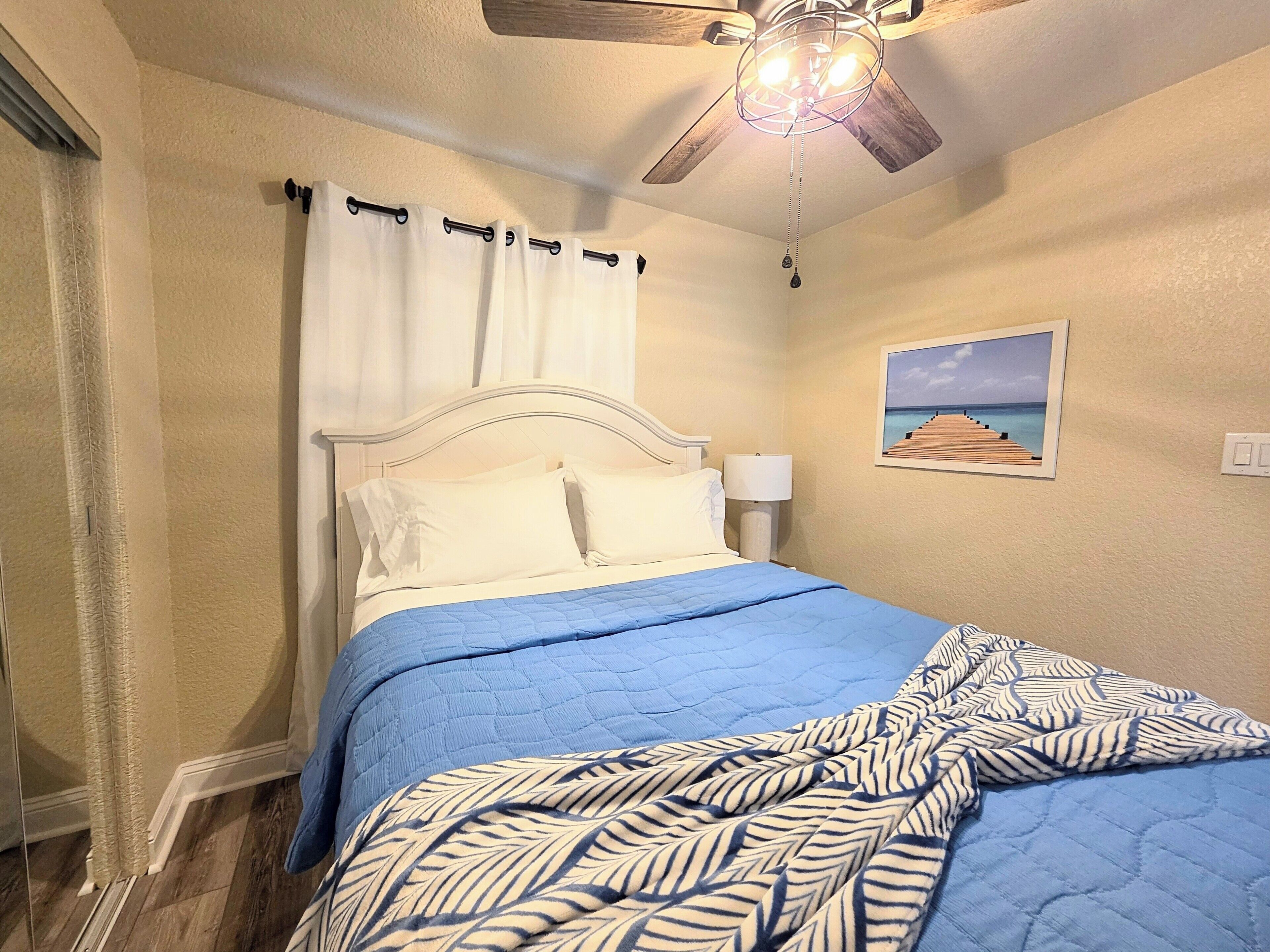 Una camera da letto con un letto, una ventola a soffitto, un comodino, una lampada e un quadro appeso al muro.