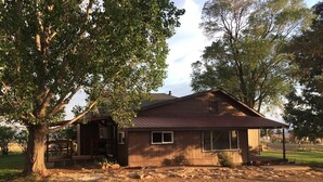 Une maison de plain-pied avec un toit en pente, un porche à l’avant et un grand arbre sur le côté gauche.