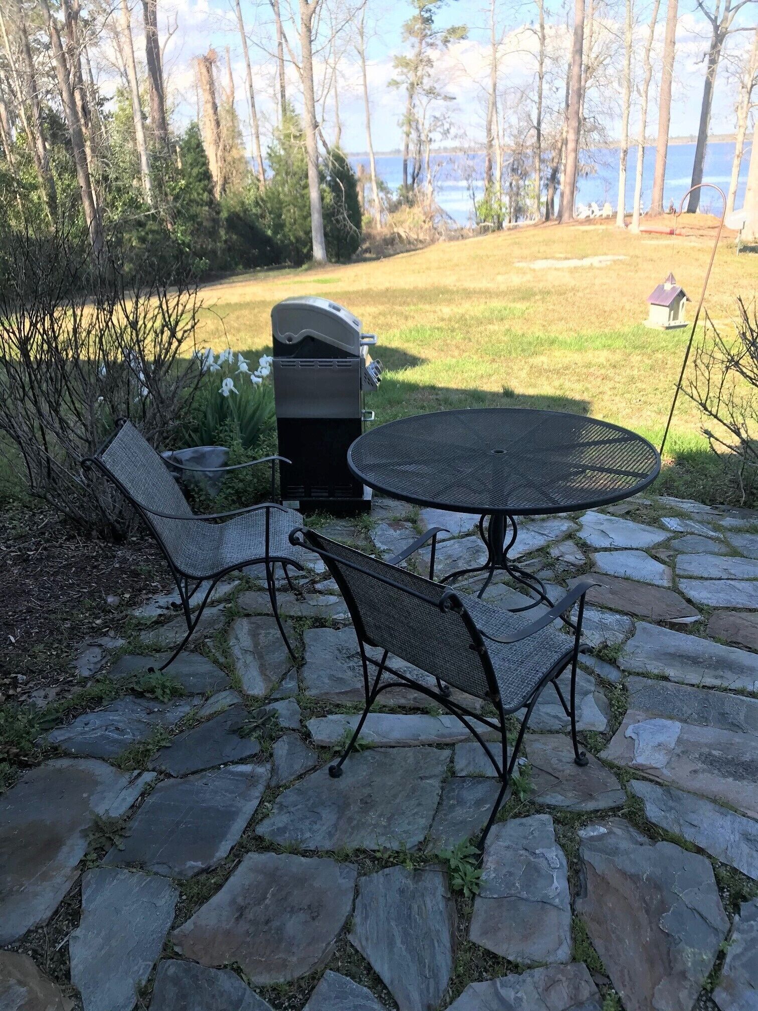 Eine Terrasse mit einem Steintisch, zwei Stühlen, einem Grill und Blick auf einen See und Bäume.