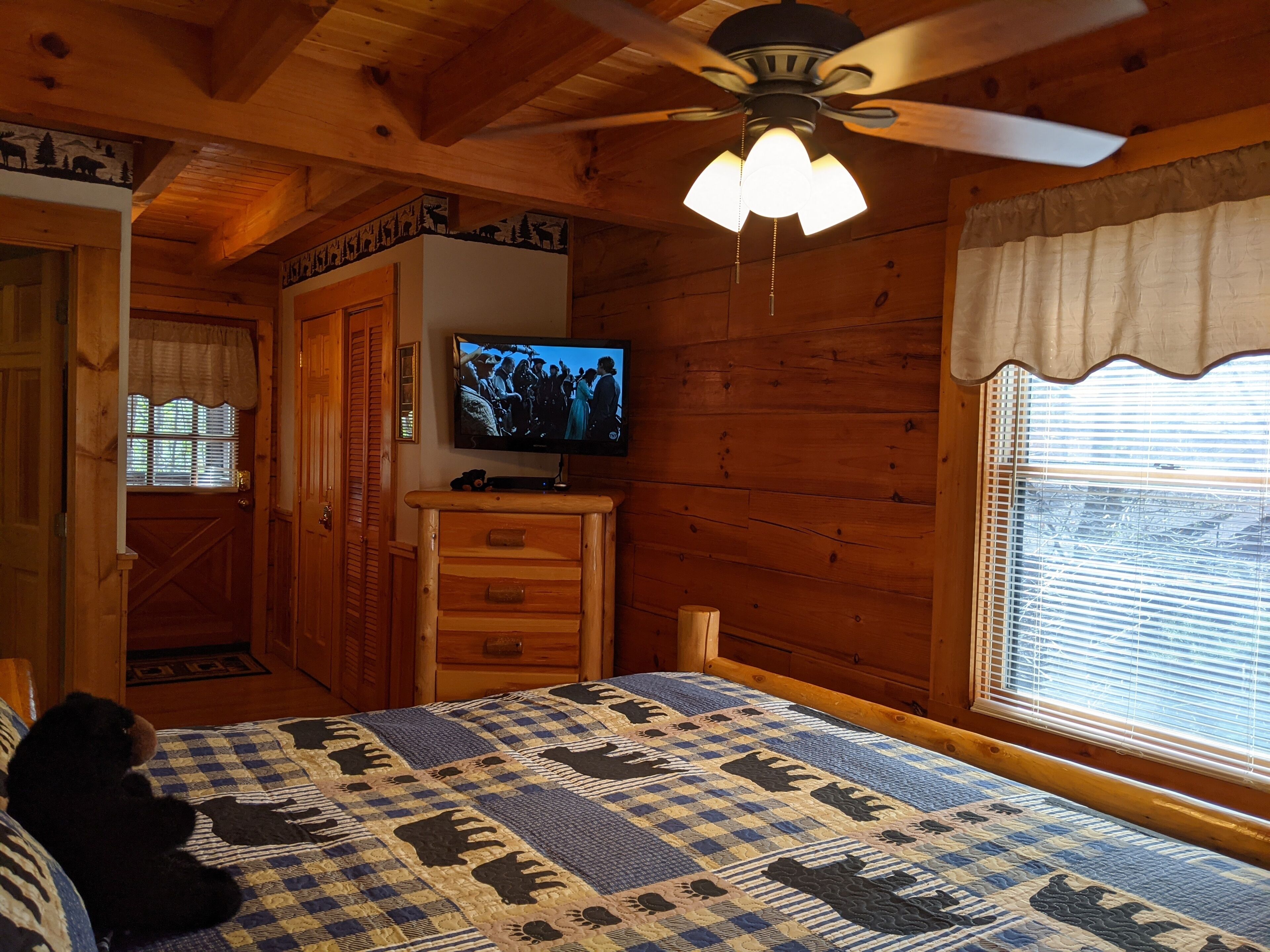Um quarto de cabana aconchegante com cama, televisão, paredes de madeira, ventilador de teto e janela com persianas.