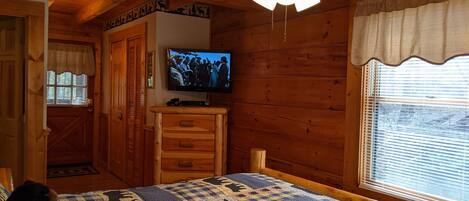 Um quarto de cabana aconchegante com cama, televisão, paredes de madeira, ventilador de teto e janela com persianas.