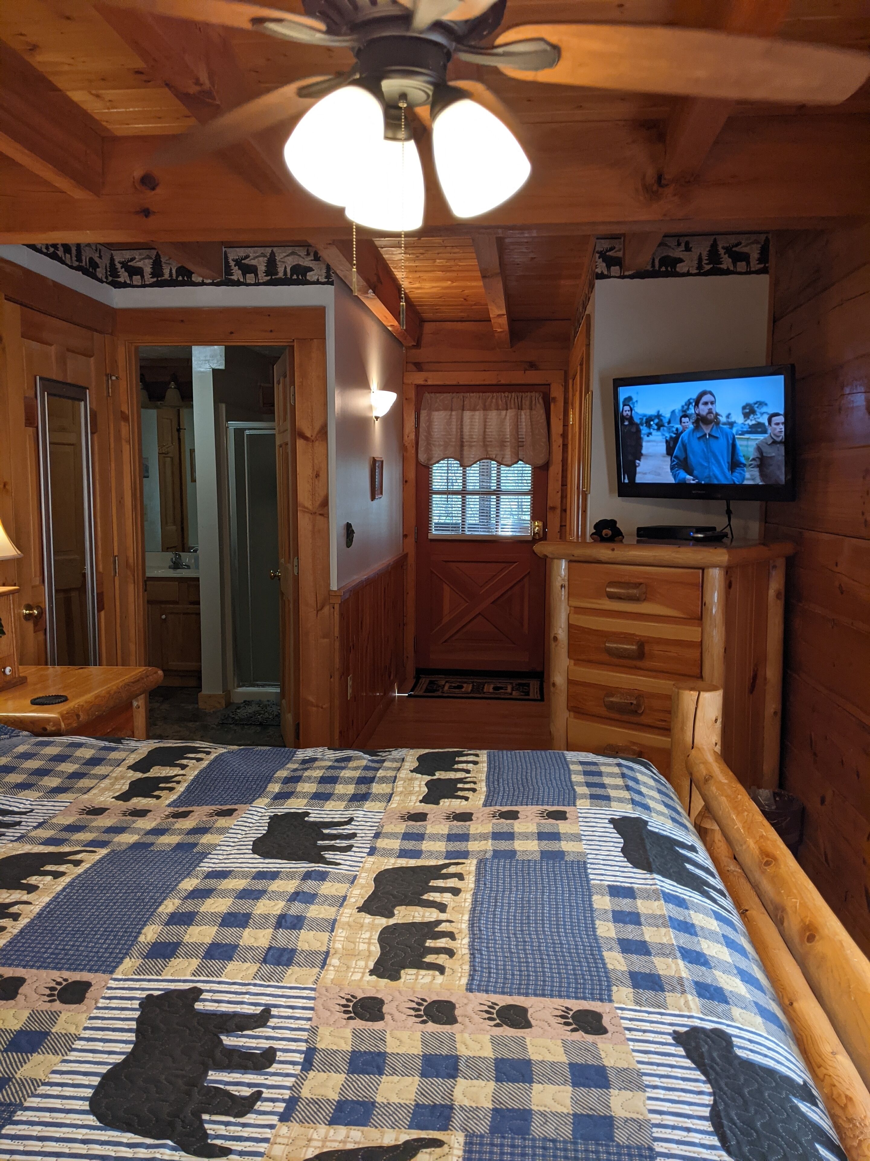 Camera in baita in legno con un letto ricoperto da una coperta a quadri con stampe di animali, una ventola a soffitto, un televisore su un supporto in legno e una porta in legno con una finestra.