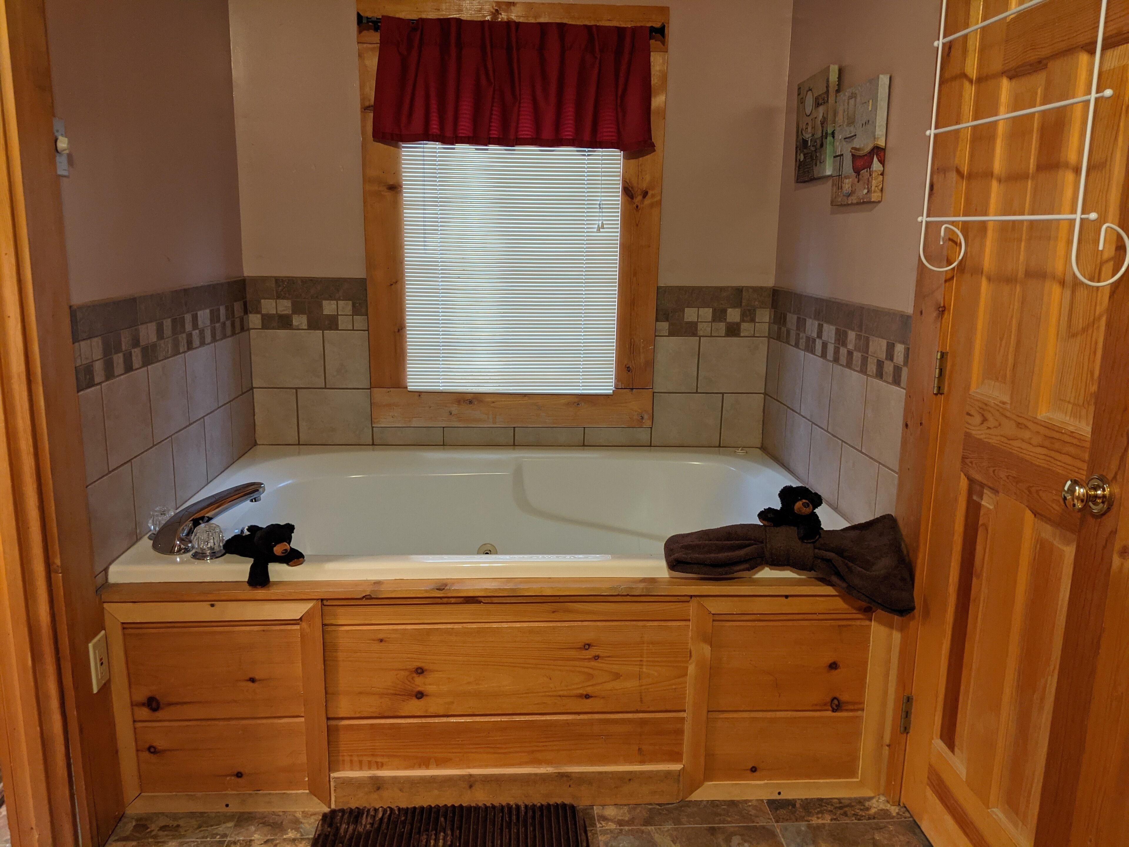 Une salle de bain avec un meuble-lavabo en bois, une baignoire, une fenêtre avec des stores et une porte dotée d’une poignée en métal.