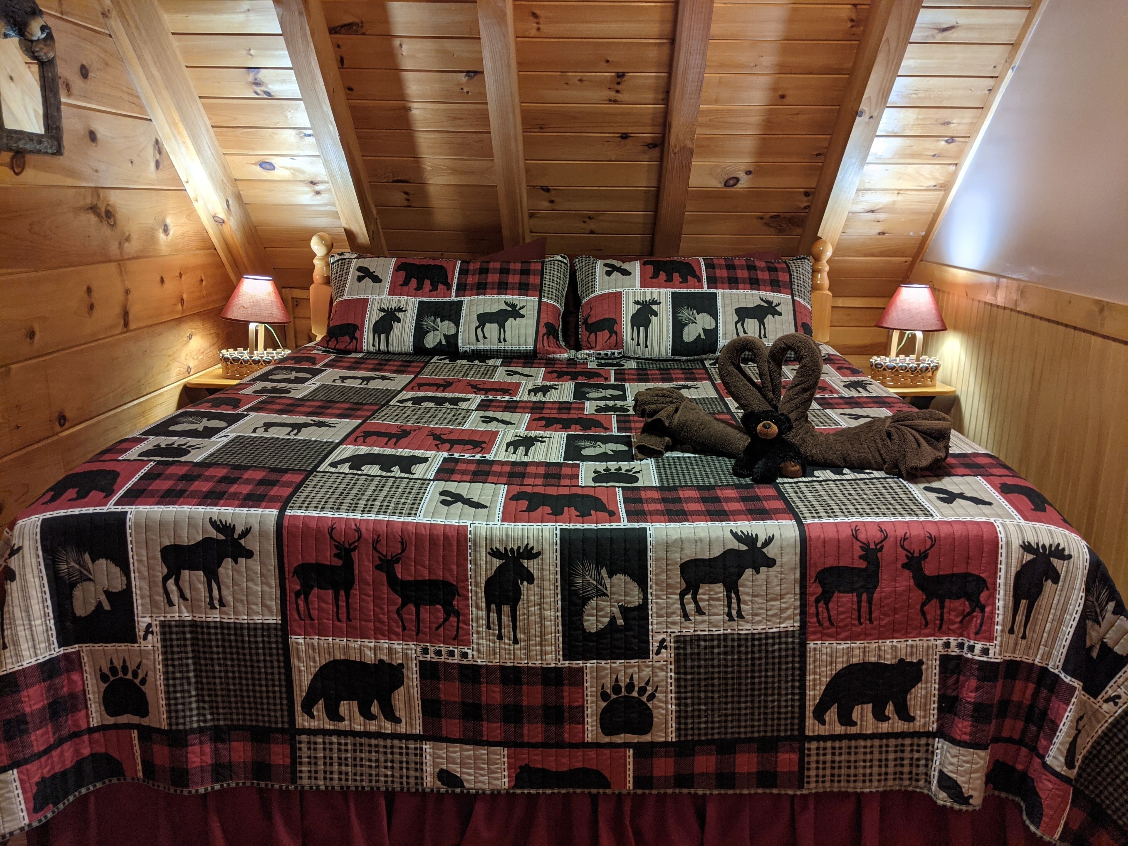 A cozy room with a wooden ceiling and walls, featuring a bed with a quilted blanket adorned with animal prints, two bedside lamps, and a teddy bear on the bed.