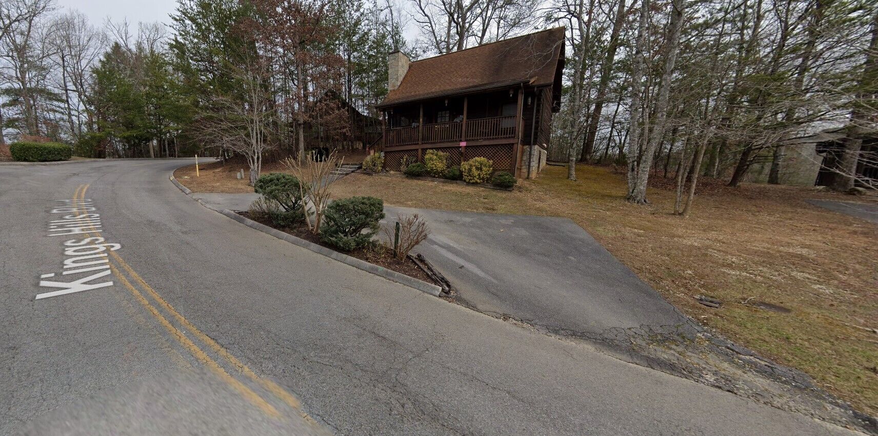 Une cabane en bois avec un porche, entourée d’arbres et bordée d’une allée.