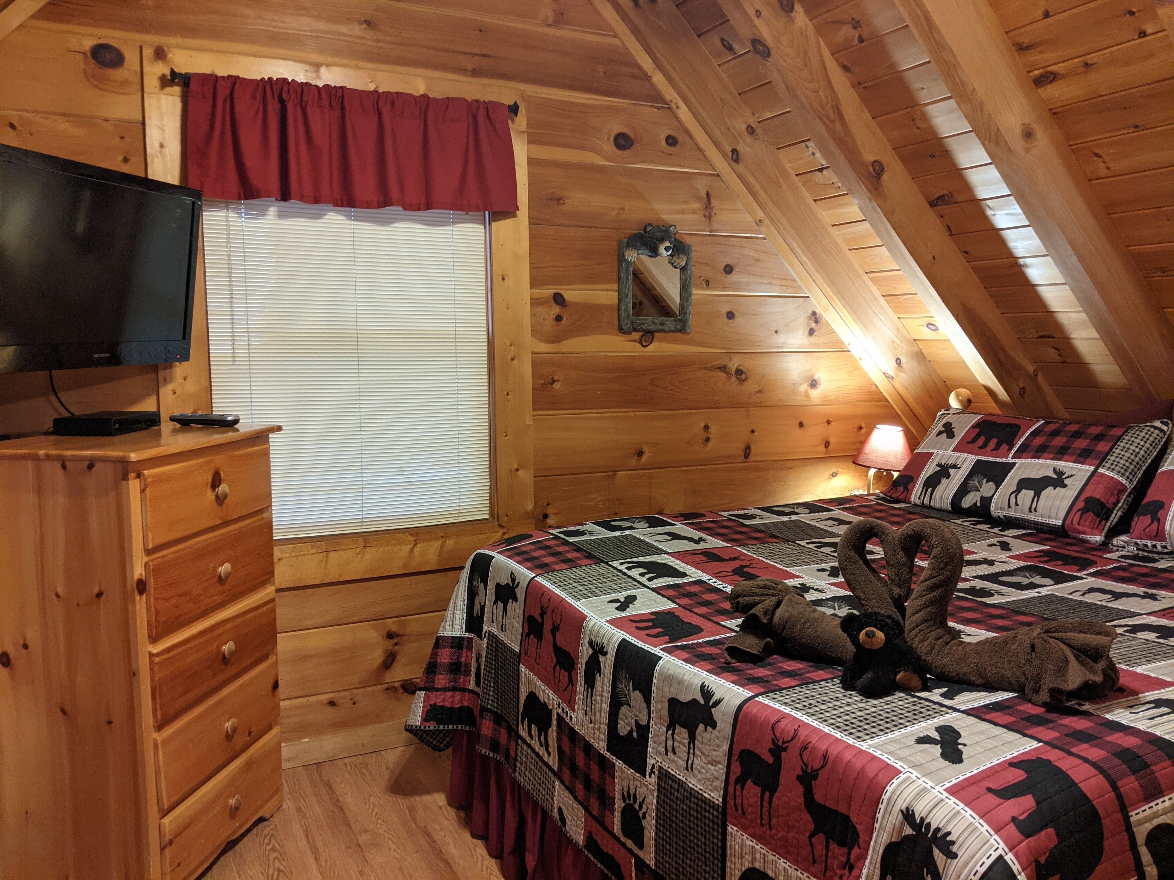 Una habitación cálida tipo cabaña con interior de madera, una cama con colcha de estampados de animales, un tocador, un televisor y una ventana con persianas.