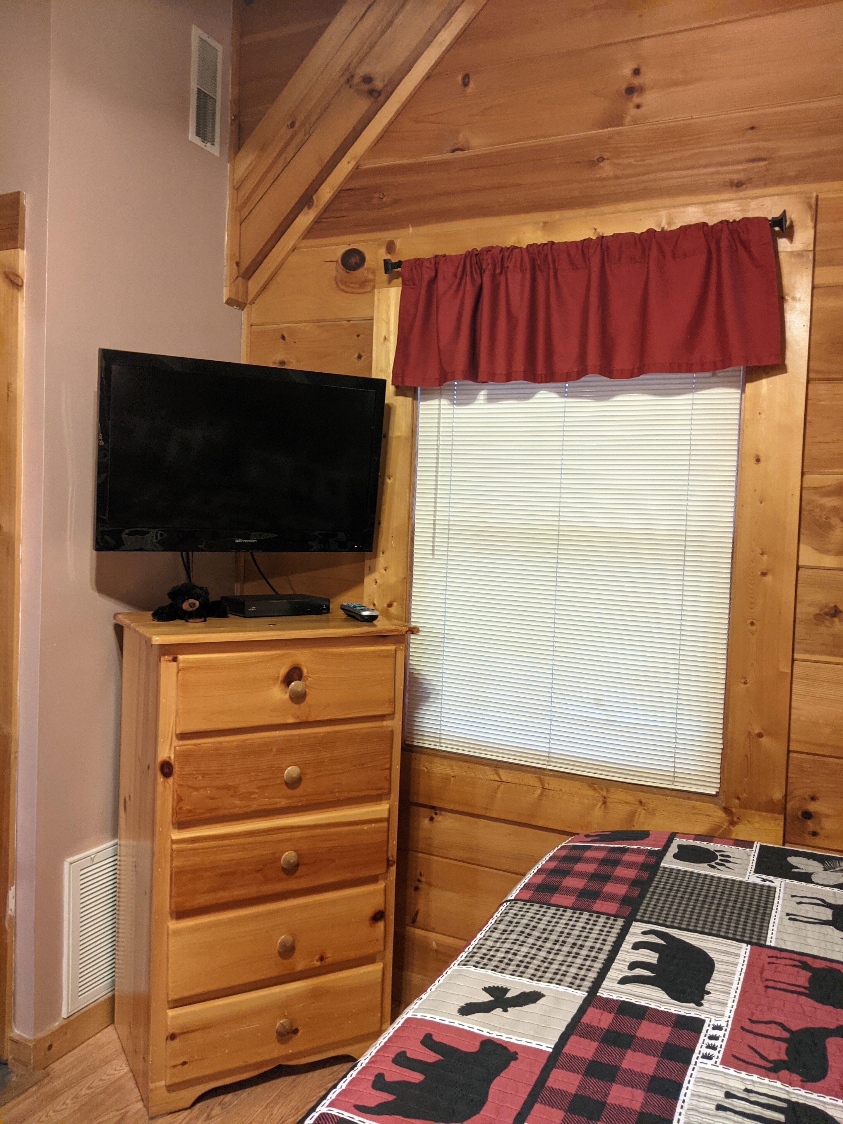 Una habitación de cabaña de madera con un televisor sobre un tocador, una ventana con persianas y un colcha con estampados de animales sobre el suelo.