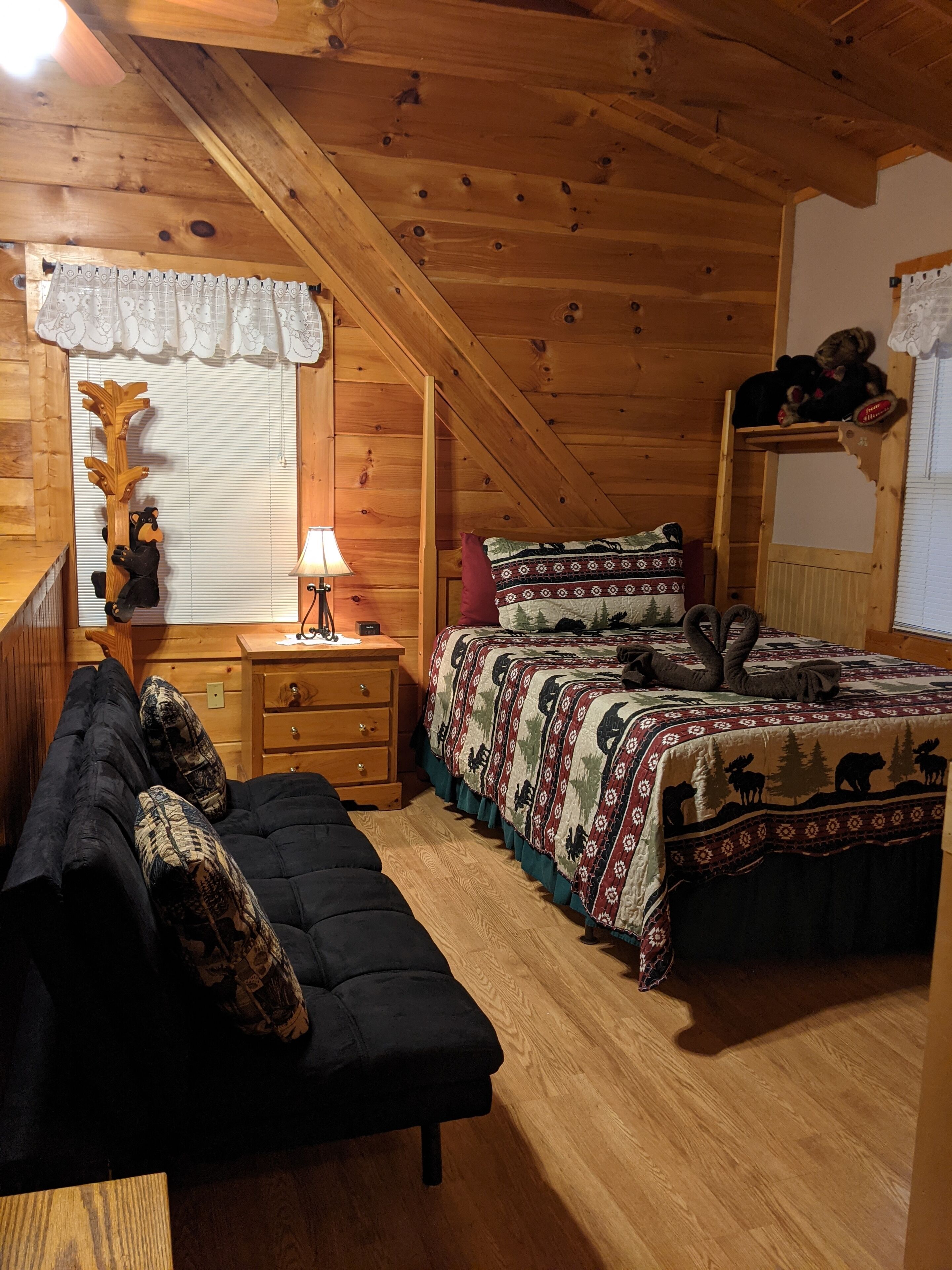 Un dormitorio de cabaña acogedor con techo de madera, una cama con manta estampada, un tocador de madera y un sofá negro.