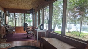 A cabin with a wooden interior, large windows overlooking a lake, a rocking chair, a table with a lantern, and a bench. - Beautiful retreat in the Adirondack Park (Malone)