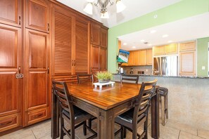A dining area with a wooden table and chairs, a potted plant, and a kitchen in the background.