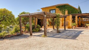 Een binnenplaats met een pergola, een geel gebouw met een pannendak en omliggend groen.
