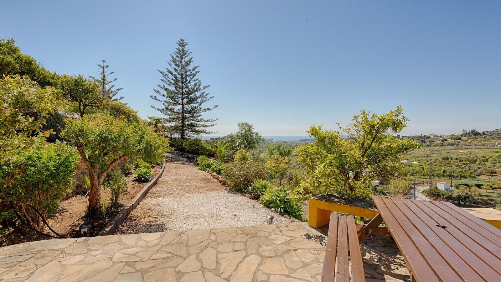 Uma trilha de pedra leva a um banco de madeira com vista para uma paisagem deslumbrante, com árvores e o mar ao longe.