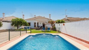 A swimming pool with a tiled deck, surrounded by a white wall and a fence. There are lounge chairs and a small garden area with a tree and potted plants. - Chalet Rocio 2 Conil de la Frontera by Ruralidays (Cádiz)