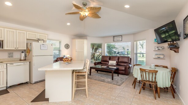 A modern kitchen with white cabinets, a central island, and a ceiling fan. There is a dining area with a table and chairs, and a living room with a brown leather sofa and a TV mounted on the wall.