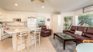 A modern kitchen with white cabinetry, a dining area with a wooden table and chairs, a brown leather sofa, and a ceiling fan.