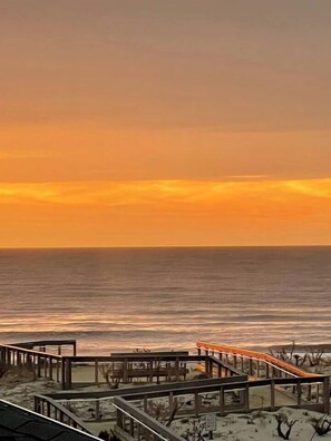 Vista à beira-mar com um calçadão de madeira que leva a uma área arenosa, com vista para o oceano.