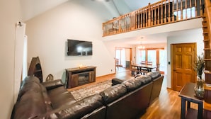 A spacious living room with a large leather sofa, a flat-screen TV mounted on the wall, and a wooden staircase leading to an upper level. - Rock Hill Retreat- a home with natural stone formations at every turn!  (Rockbridge)