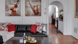 A living room with a black leather sofa, red and floral cushions, a coffee table with a bottle and decorative items, and two wall art pieces depicting musical instruments. - Downtown Memphis 2 BR Eleanor Rigby suite Apt 101 (Memphis)