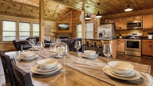 Interior de una cabaña de madera con una mesa de comedor dispuesta para seis personas, zona de cocina con electrodomésticos modernos y un rincón de estar acogedor.