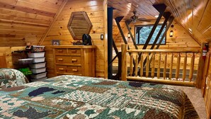 A cozy cabin bedroom with a large bed, wooden walls, a dresser, and a window with a view.
