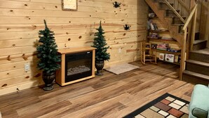 A log cabin interior with a fireplace, two Christmas trees, a wooden staircase, and a bookshelf with books and games.