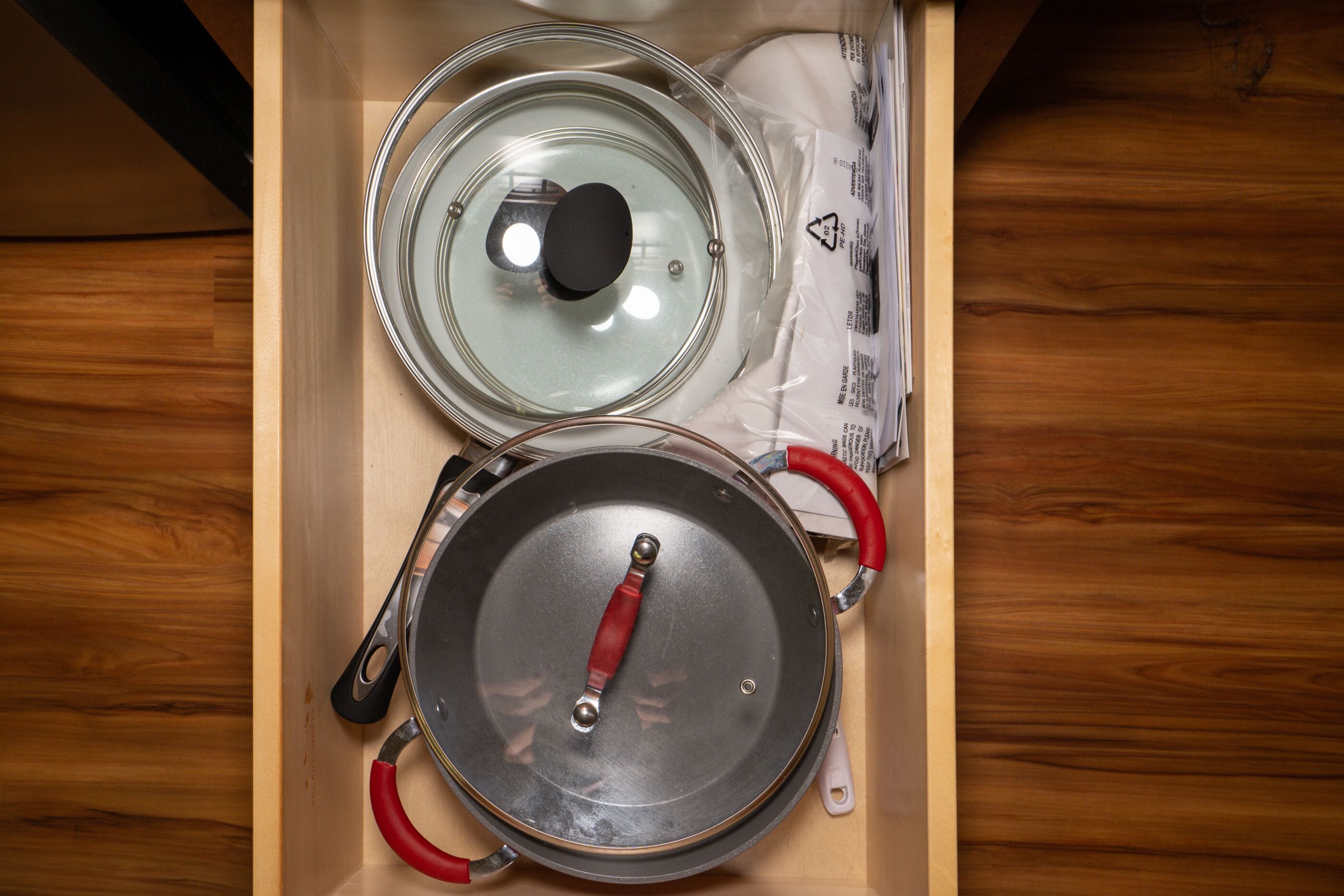 Un cassetto della cucina con una padella con manico rosso e un coperchio in vetro.