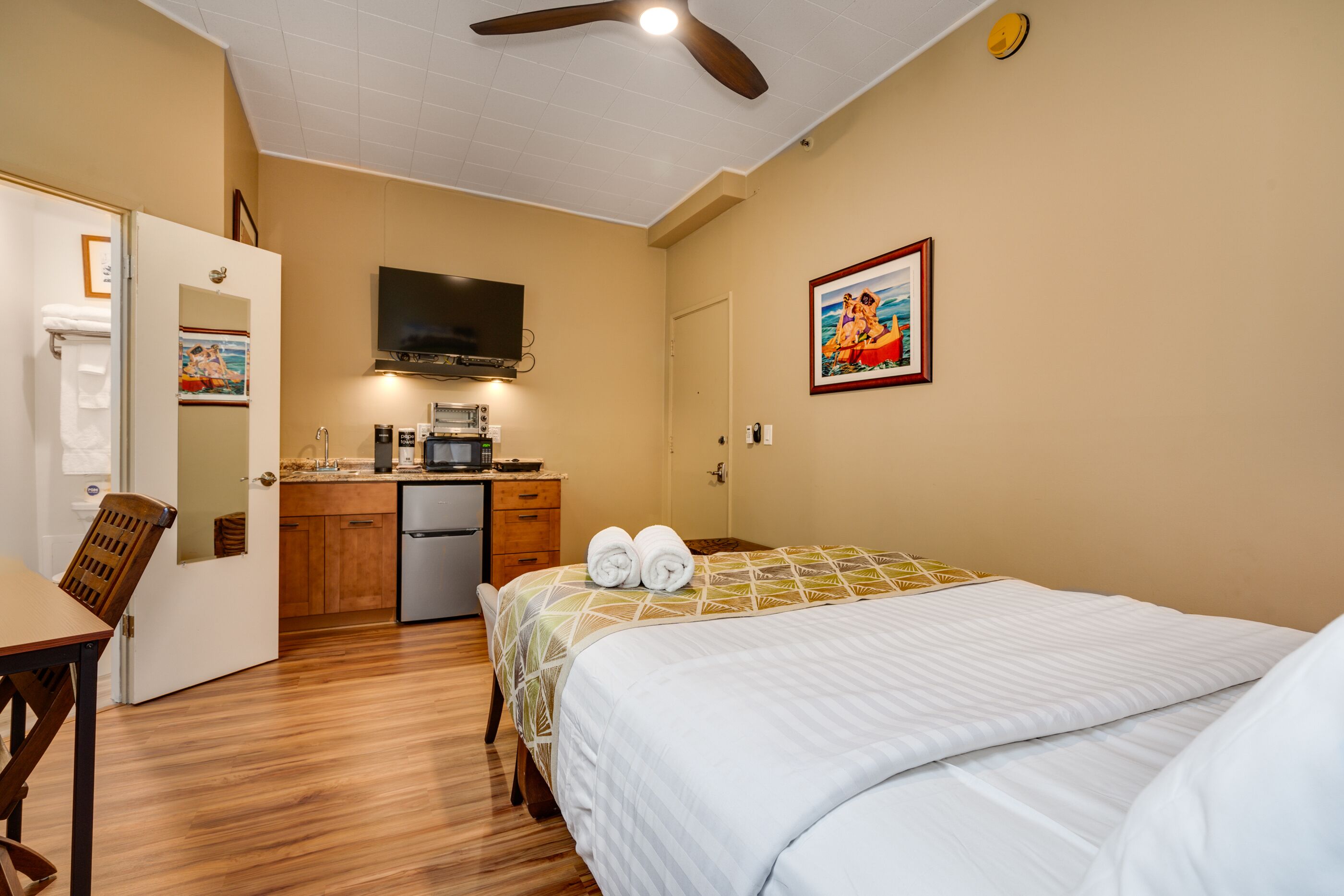 Een hotelkamer met een bed, een bureau, een kitchenette en een plafondventilator.