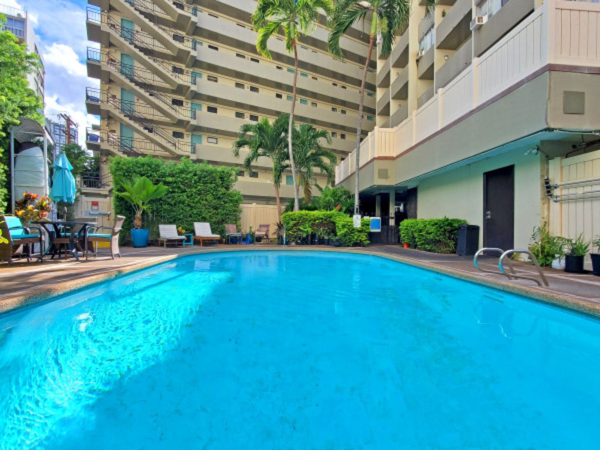 A swimming pool surrounded by lounge chairs and potted plants, with a multi-story building in the background.