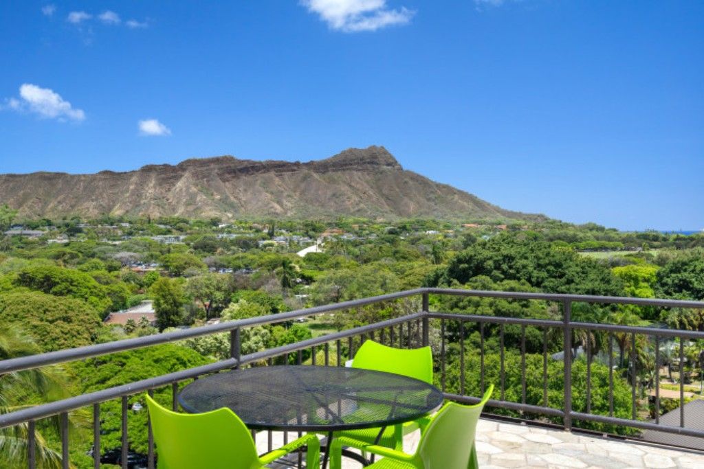 A balcony with a round table and green chairs, overlooking a mountainous landscape and a lush green valley.