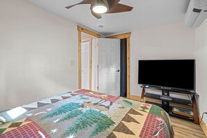 Un dormitorio con cama, televisor sobre un soporte, ventilador de techo y una puerta.