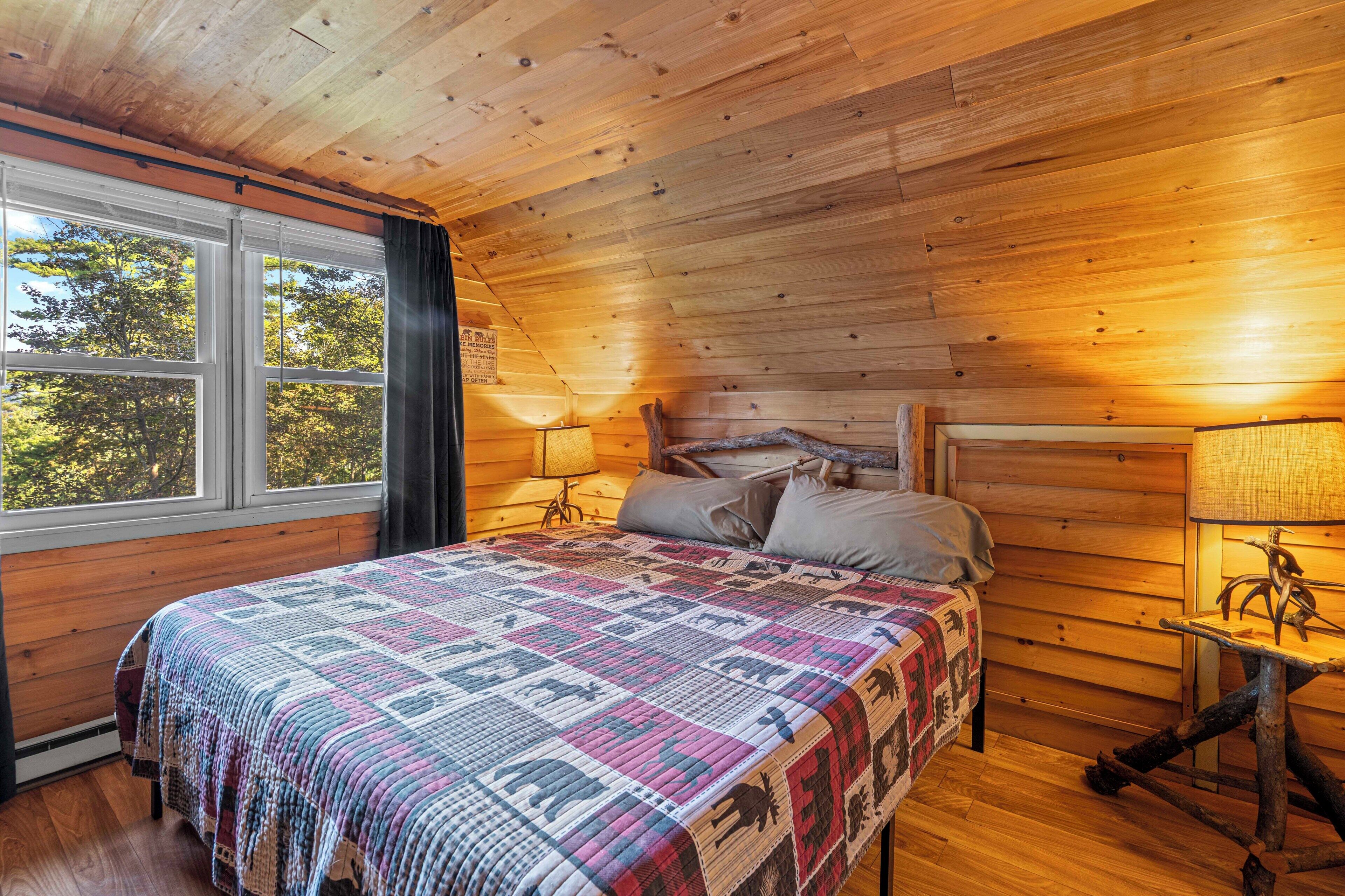 A wooden cabin bedroom with a bed, two bedside lamps, a window, and a view of trees.