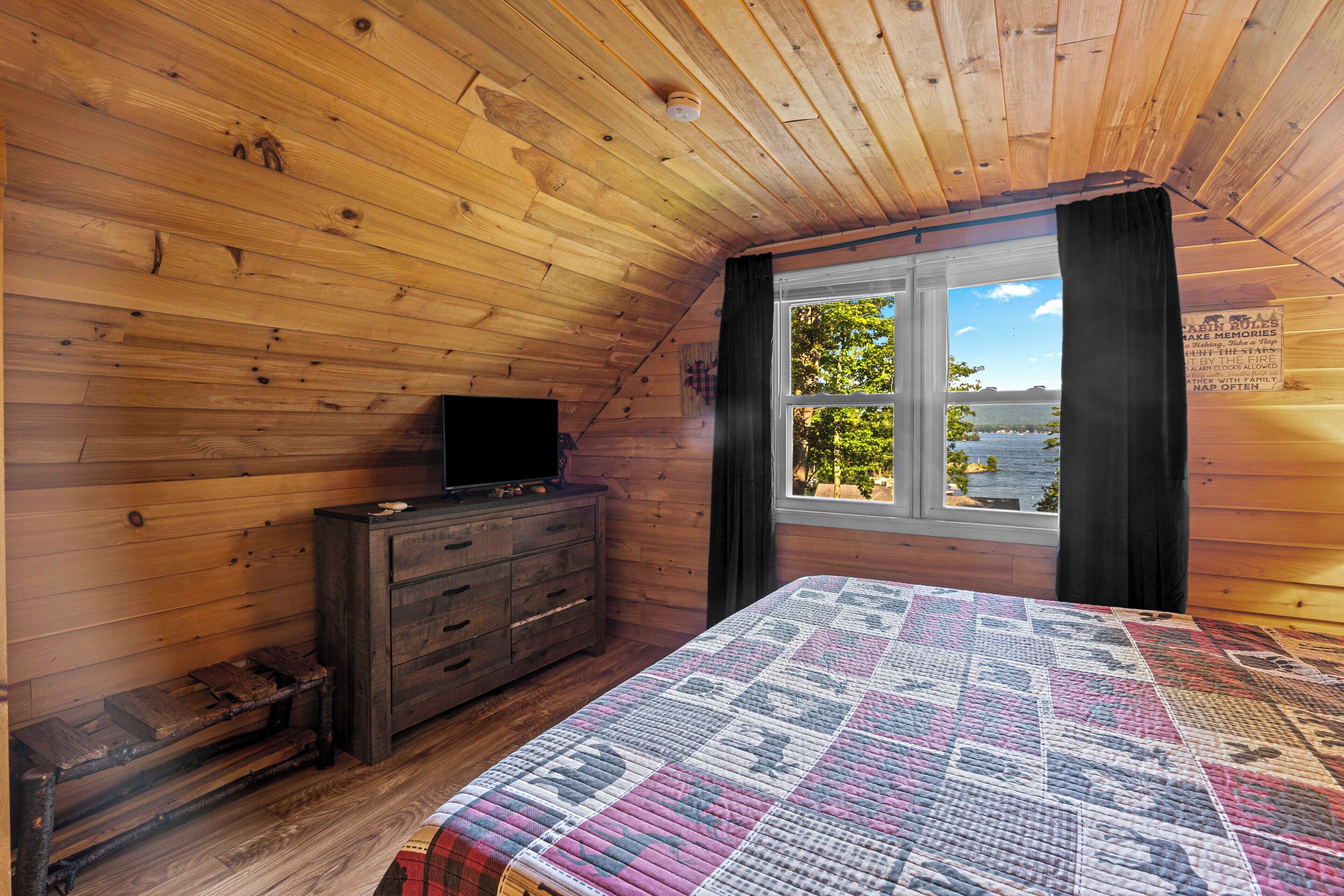 Quarto de cabana com teto de madeira, uma cama com colcha, cômoda e janela com vista para árvores e água.