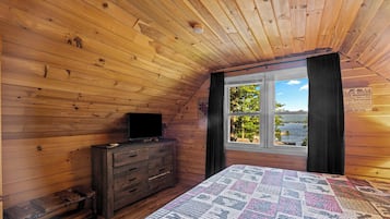 Quarto de cabana com teto de madeira, uma cama com colcha, cômoda e janela com vista para árvores e água.