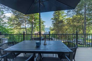 A wooden deck with a table and chairs, overlooking a body of water and trees. - Lake George Chalet • Beach Access • Fire Pit • Sleeps 8• Pet friendly (Lake George)
