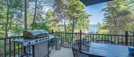 An outdoor deck with a barbecue grill, a table, and chairs, overlooking a forest and a body of water.