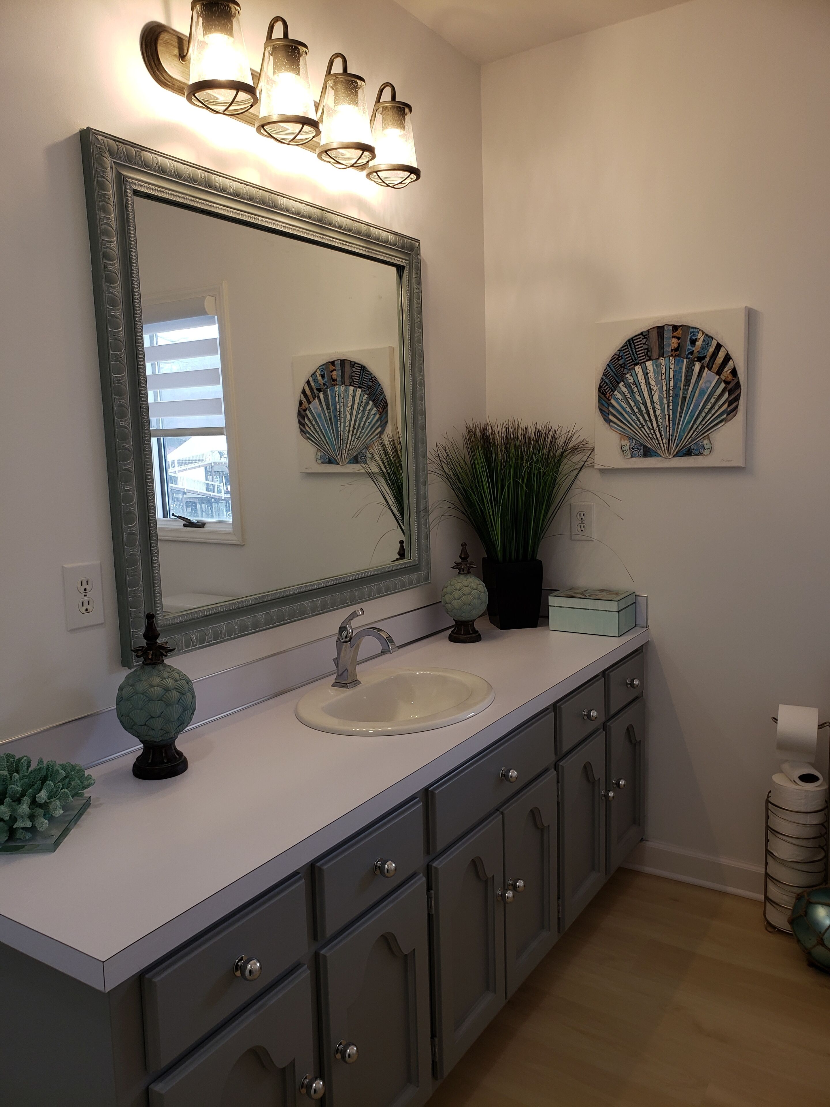 Un baño con un espejo grande, un lavabo y un mueble de tocador con cajones. Hay objetos decorativos en la encimera y una planta en maceta.